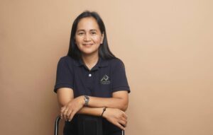 Portrait of a smiling woman in a navy polo leaning on the back of a chair against a beige backdrop, arms resting softly in front.