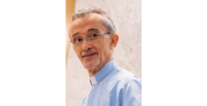 Portrait of an older man with glasses wearing a light blue clerical shirt and white collar, smiling.