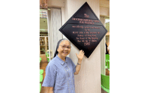 Smiling nun in a blue shirt stands beside a diamond-shaped plaque commemorating the opening of the Diocesan Pastoral Center for the Philippines (January 4, 1987).