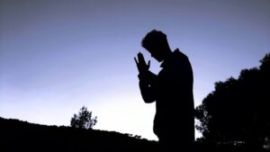 Silhouette of a person standing with hands pressed together in prayer at sunset.
