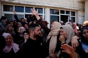 Crowd of grieving people, many women wearing headscarves, gathering outside a building with raised hands and somber expressions.