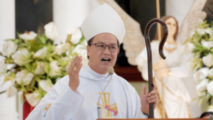 Catholic bishop in white vestments delivering a sermon at the altar, holding a shepherd's crook and speaking into a microphone