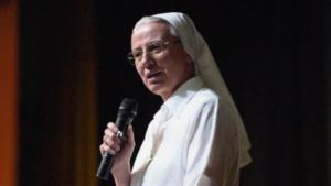 A nun in a white habit speaks into a handheld microphone on stage.