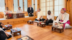 Diverse group of men in religious and formal attire sit on a wooden floor and raised platform, sharing tea in a sunlit room.